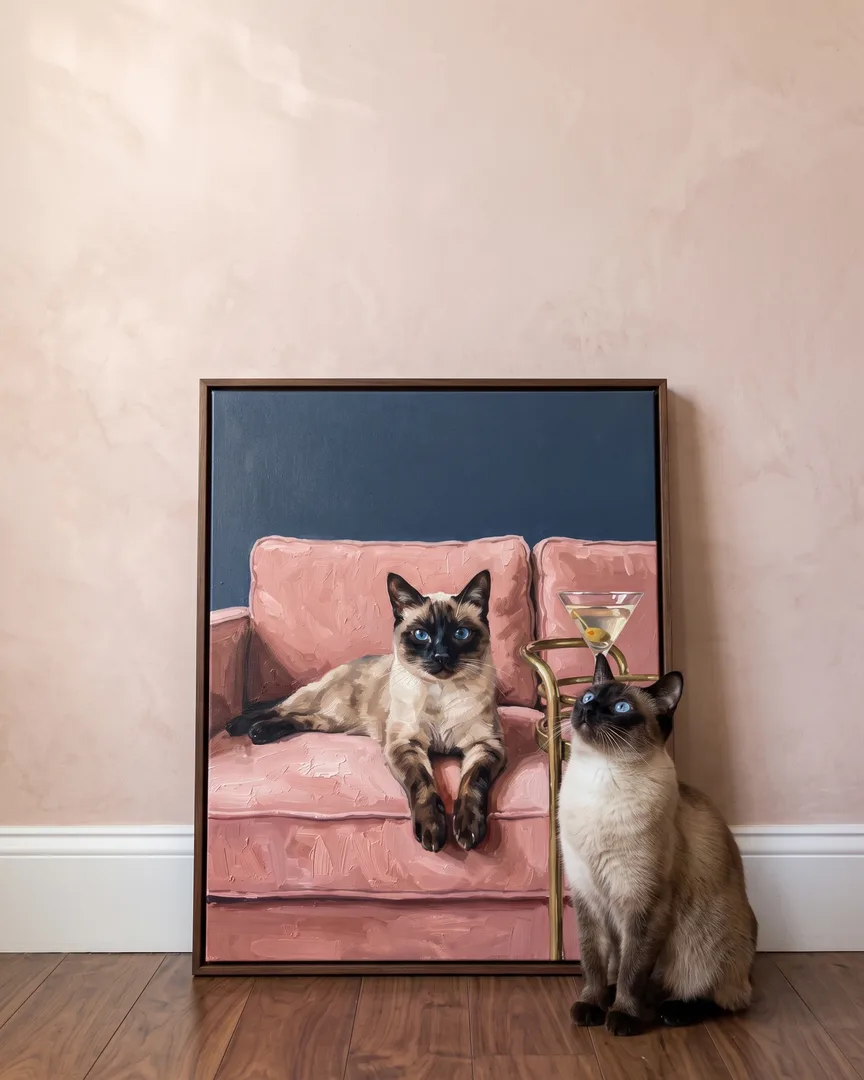 Sable, a Siamese, beside a brown framed canvas of their own Olive & Velvet portrait.
