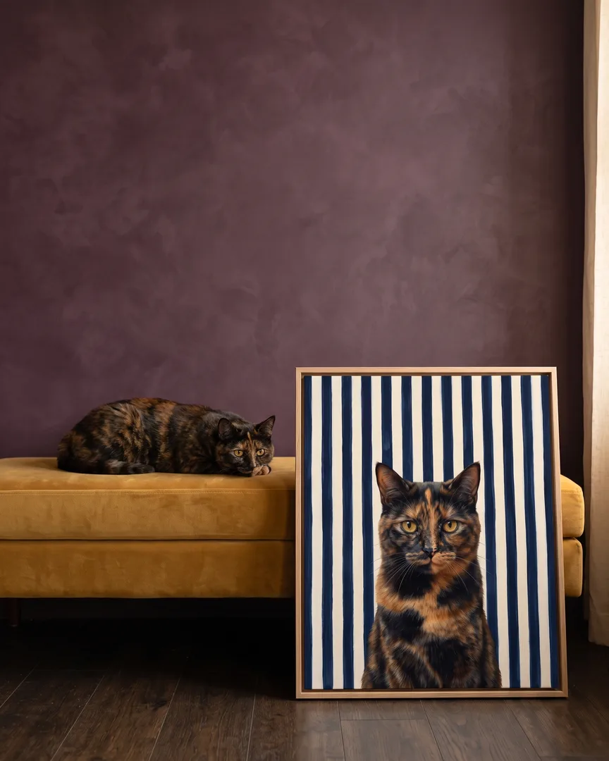 Autumn, a tortoiseshell domestic shorthair, beside a natural-oak framed canvas of their own Riviera Stripe portrait.