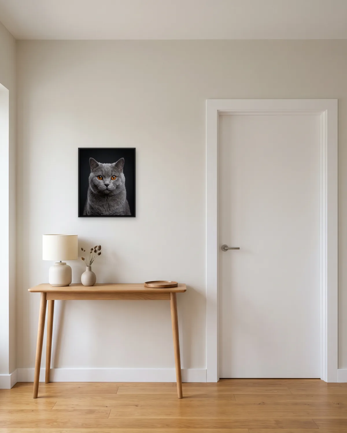 A 16 x 20 in framed canvas of a grey cat in a thin black float frame, hung above a wooden console in a hallway beside a white door